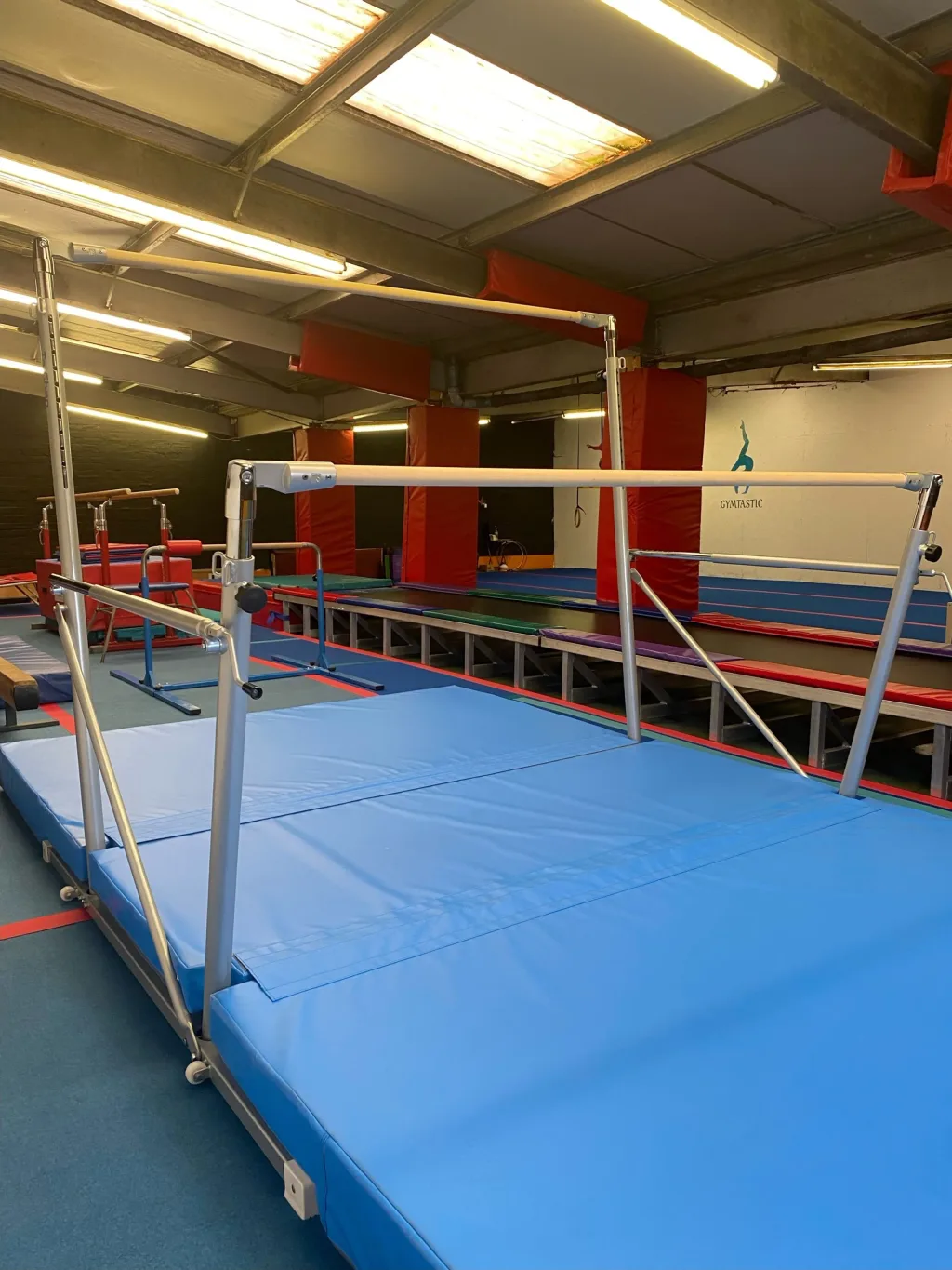 Freestanding uneven bars with wooden rails on a silver frame above a blue foldable mat in a gym, wall text 'GYM TASTIC'.