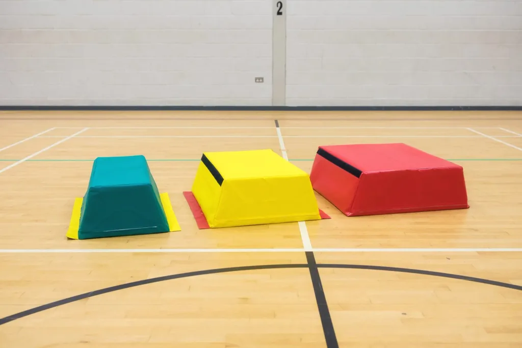 Three PVC-coated nylon vaulting boxes in teal, yellow, and red on a wooden gym floor with wall marking 2.