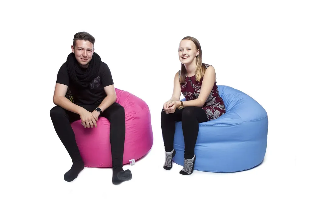 Two teenagers or young adults sitting on cotton bean bags, one pink and one sky blue, smiling against a white background.