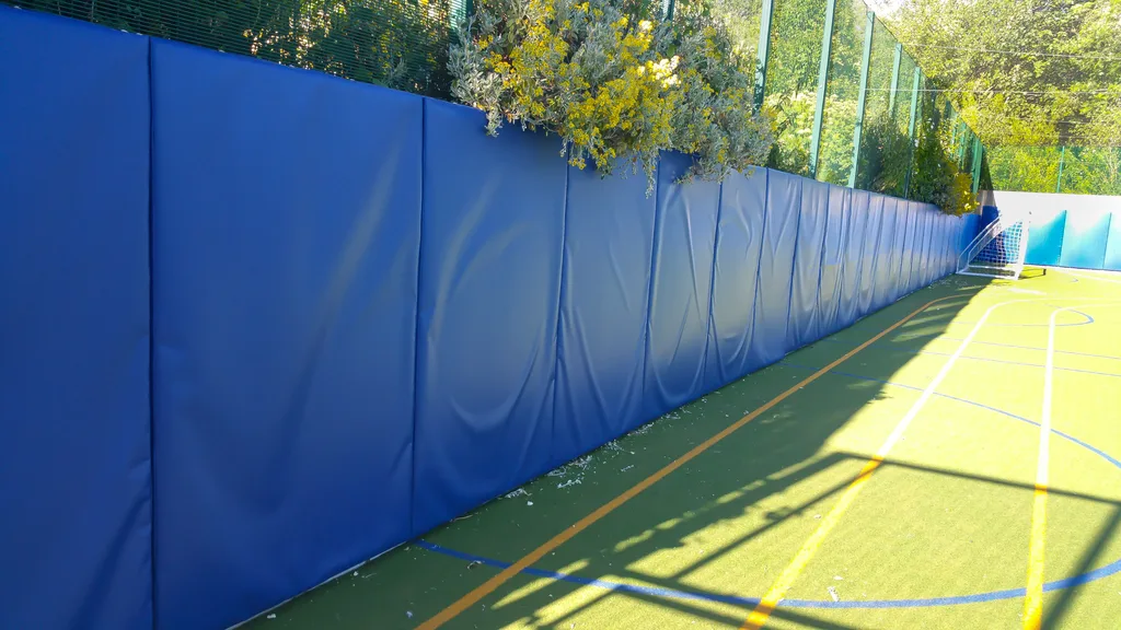 Blue outdoor safety padding panels line a fence beside a green sports court with yellow and blue markings and a small goal at the far end.