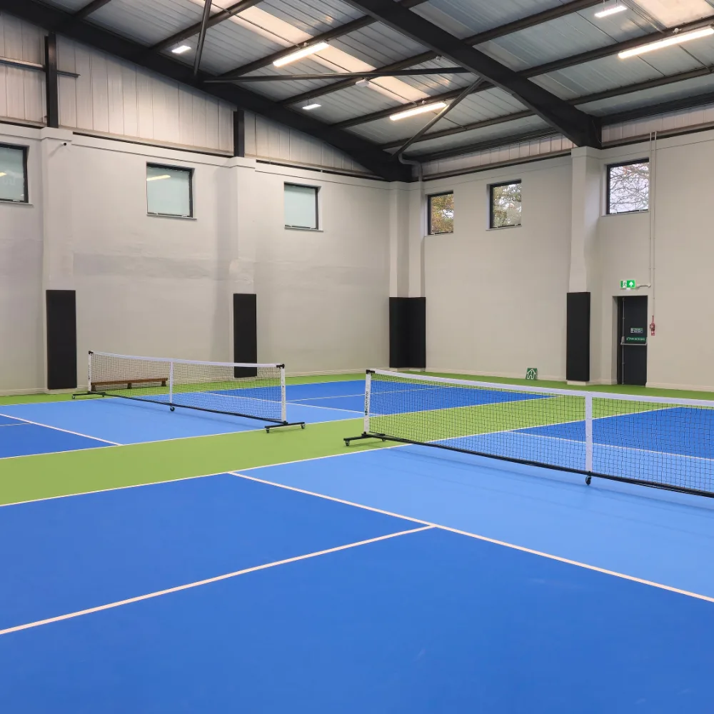 Indoor pickleball courts with blue playing surfaces, green borders, two portable nets, and tall black PVC-covered wall padding along pale walls.