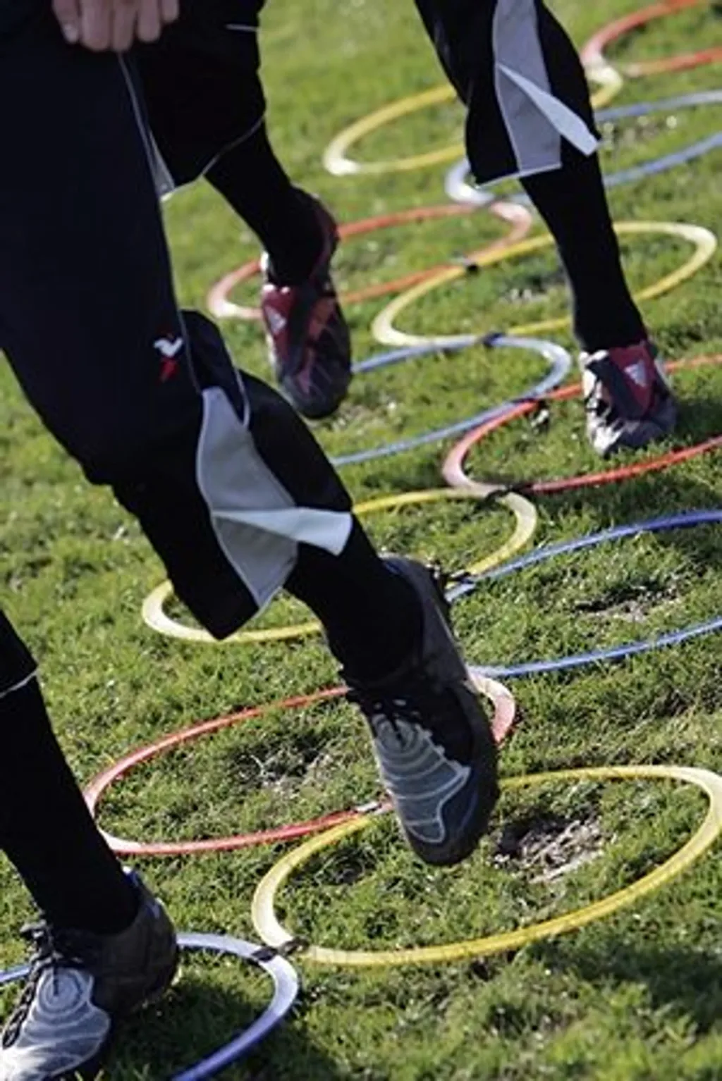 Athletes' lower legs and running shoes moving through 12 interlocked red, yellow, and blue agility hoops on grass.
