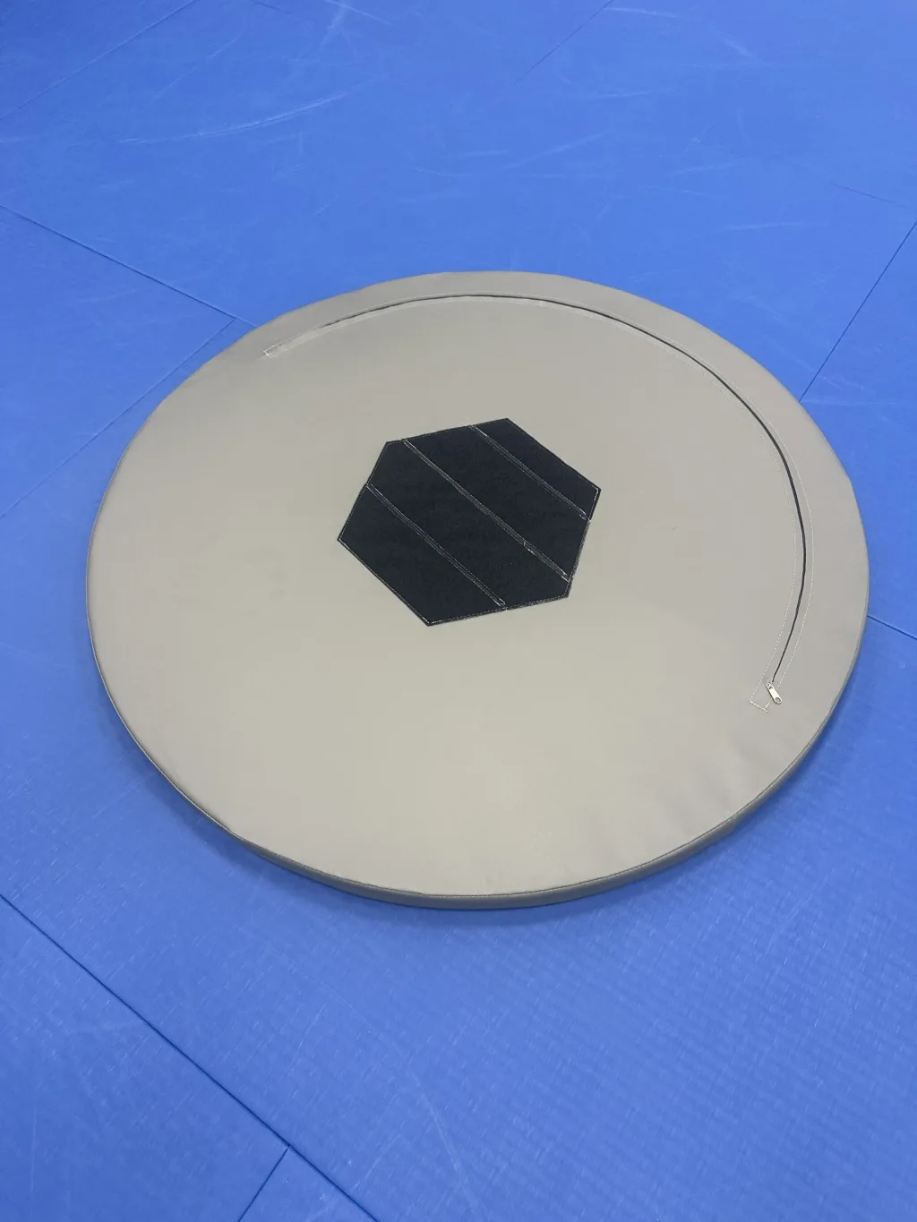 Round beige PVC-covered foam training table with a central black hexagonal hook-and-loop patch resting on blue floor mats.