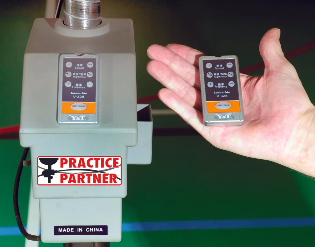 Badminton shuttle feeder with a hand holding the remote; label reads PRACTICE PARTNER and MADE IN CHINA, remote shows V-328.