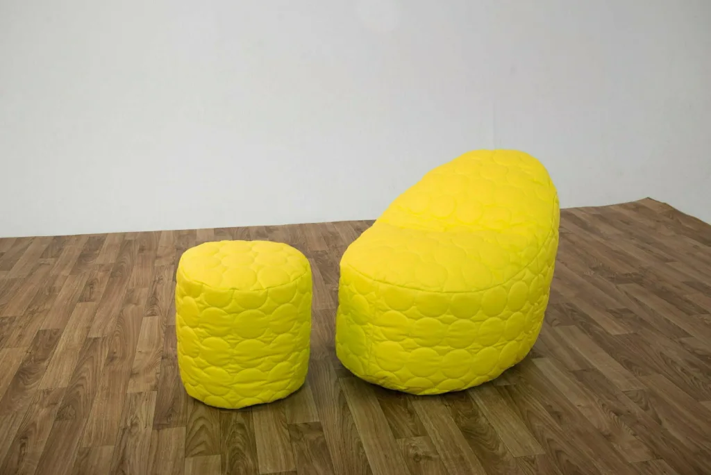 Yellow padded child-sized bean chair with back support and matching round stool on a wood floor against a plain pale wall.