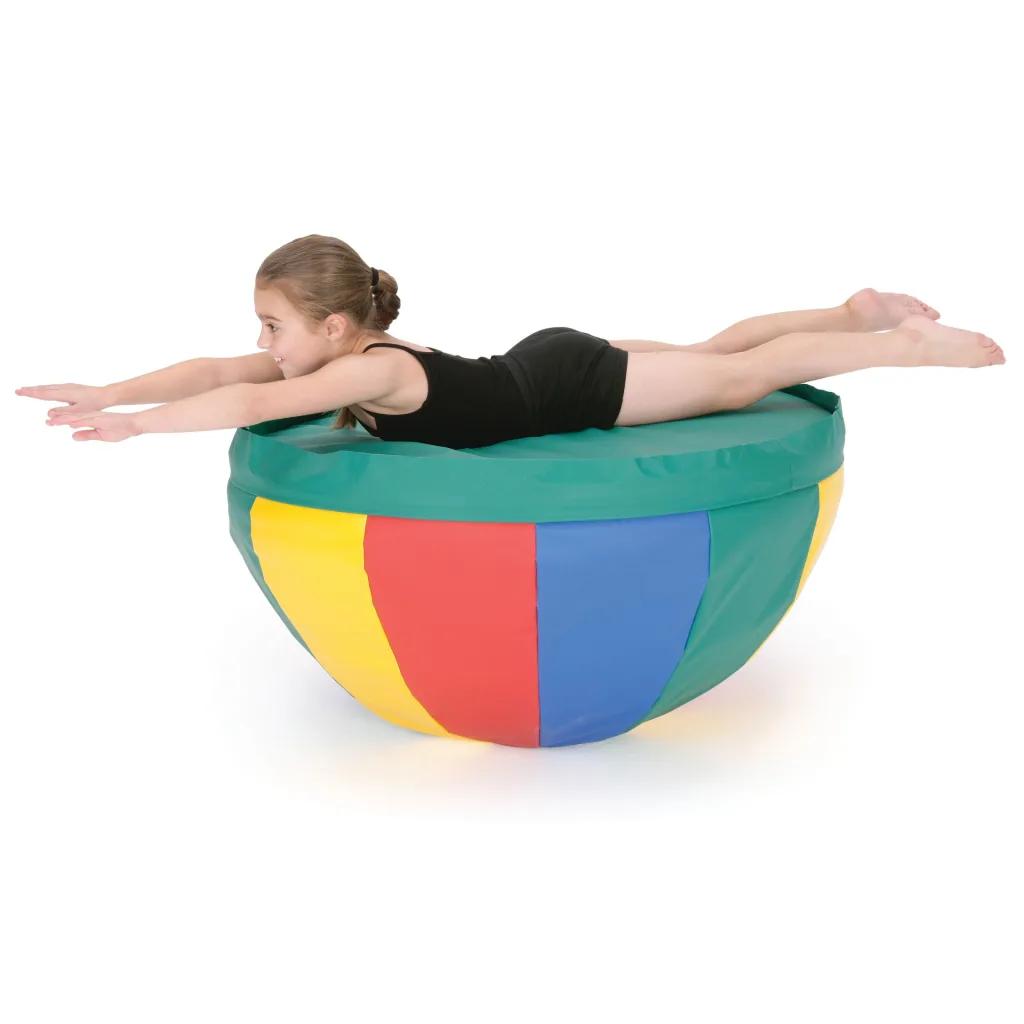 Young girl in a black leotard lying on her stomach with arms extended on a green-topped, multicolored round soft play cushion.