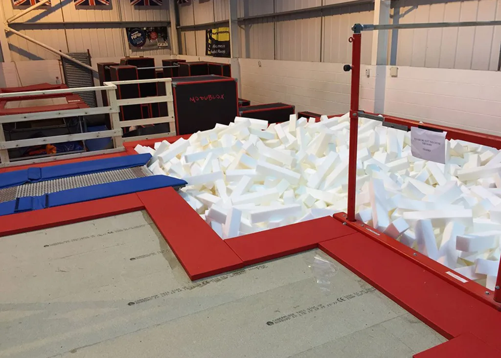 Foam pit filled with white rectangular foam blocks beside red-padded edges and two blue trampoline beds in an indoor gym.