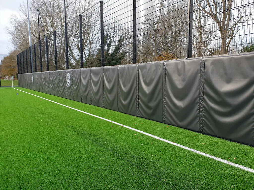 Padded gray protective panels line a black mesh fence beside a green artificial turf field with a white sideline.