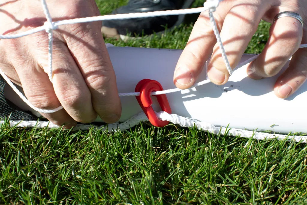 Hands fastening a white goal net to a white tubular frame using a red plastic clip on green grass.