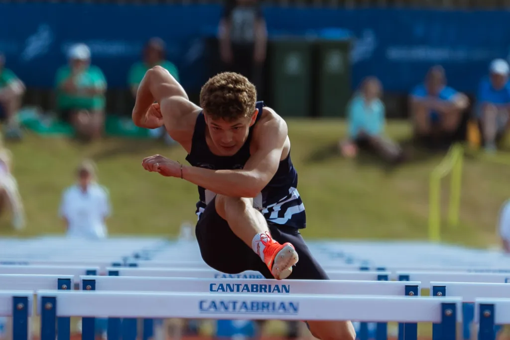 Male hurdler clearing a white steel hurdle labeled CANTABRIAN while wearing a navy singlet and bright orange spikes, spectators blurred behind.