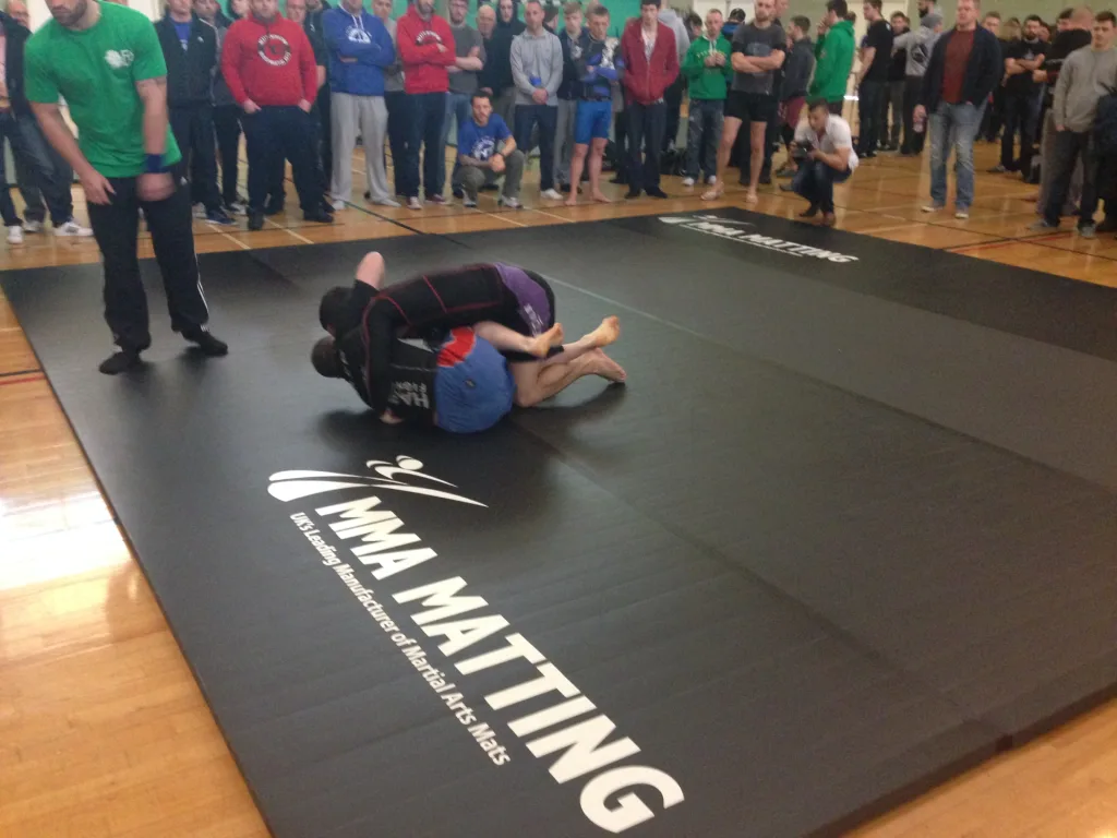 Two grapplers in rashguards wrestling on a black roll-out martial arts mat while a crowd watches in a gymnasium.