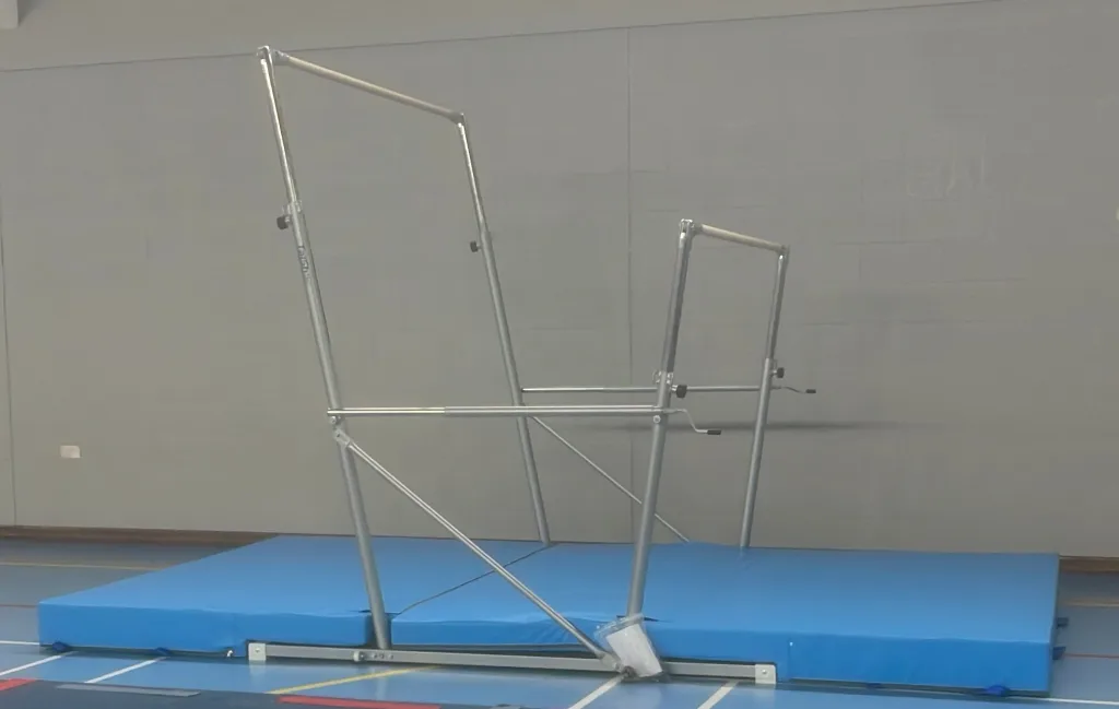 Freestanding uneven bars with two adjustable silver frames and wooden bars mounted over a blue foldable landing mat in a gym.
