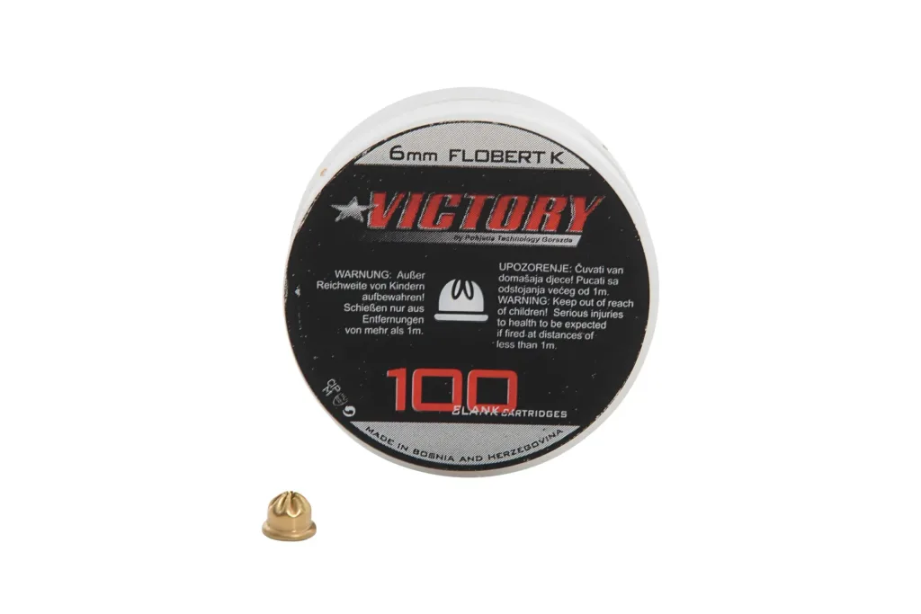 Round tin labeled 6mm FLOBERT K and 100 BLANK CARTRIDGES with a single crimped brass .22 (6mm) blank cartridge in front.