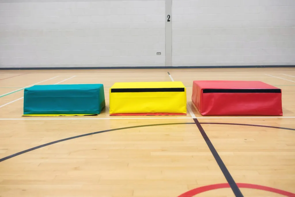 Three flame-retardant PVC-coated nylon foam vaulting boxes in green, yellow, and red on a gym floor, wall marked 2.