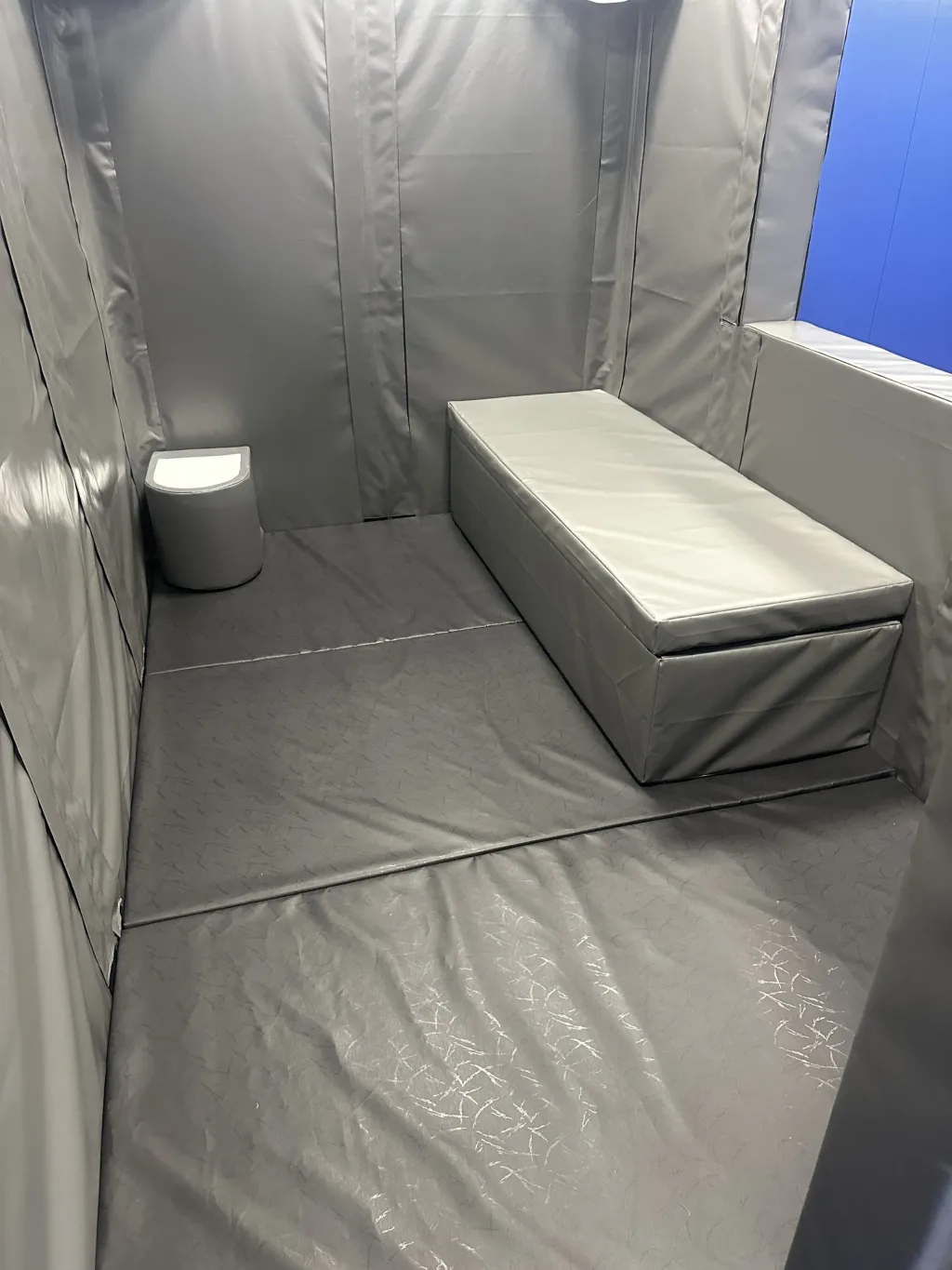 Foam bed with removable mattress wrapped in grey heavy-duty PVC against padded walls in a custody cell training setup, with a small padded toilet.