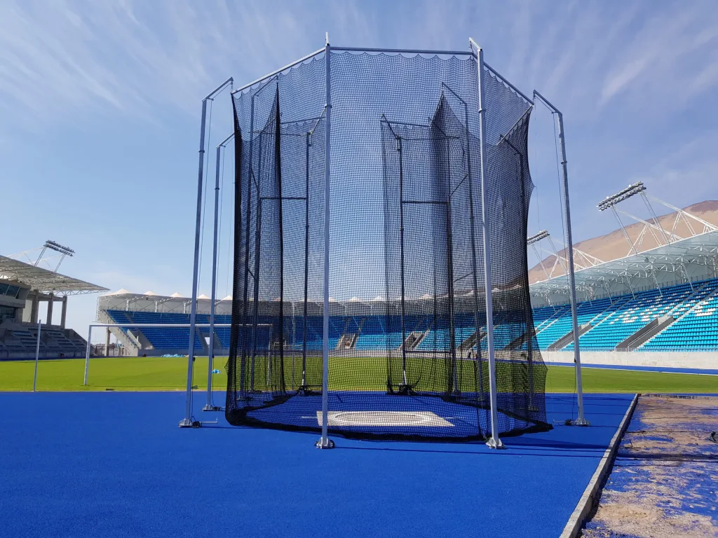 Steel hammer and discus cage with black netting and galvanised steel pillars on a blue synthetic infield in an empty stadium.