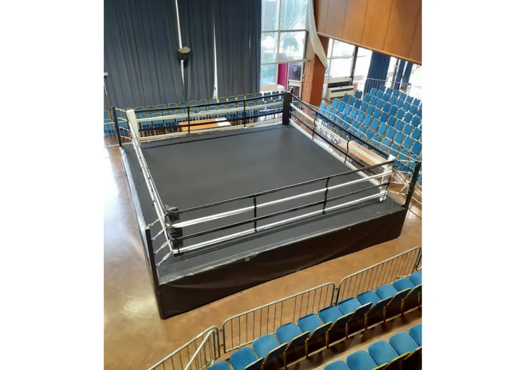 Competition ring with raised deck, steel powder-coated frame, black canvas and skirts, corner posts with three ropes, and blue spectator seats.