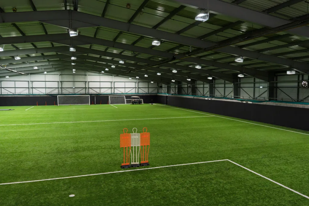 Indoor sports pitch with green artificial turf, perimeter safety padding along the walls, three training mannequins in the foreground and goals at the far end.