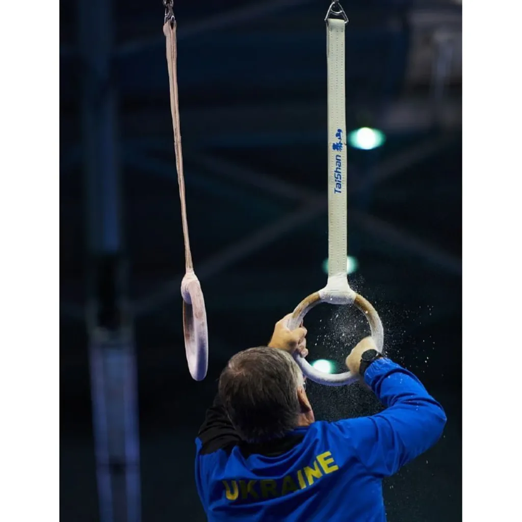 Person in blue jacket labeled "UKRAINE" seen from behind gripping a gymnastics ring as chalk dust sprays, with a second ring hanging nearby.