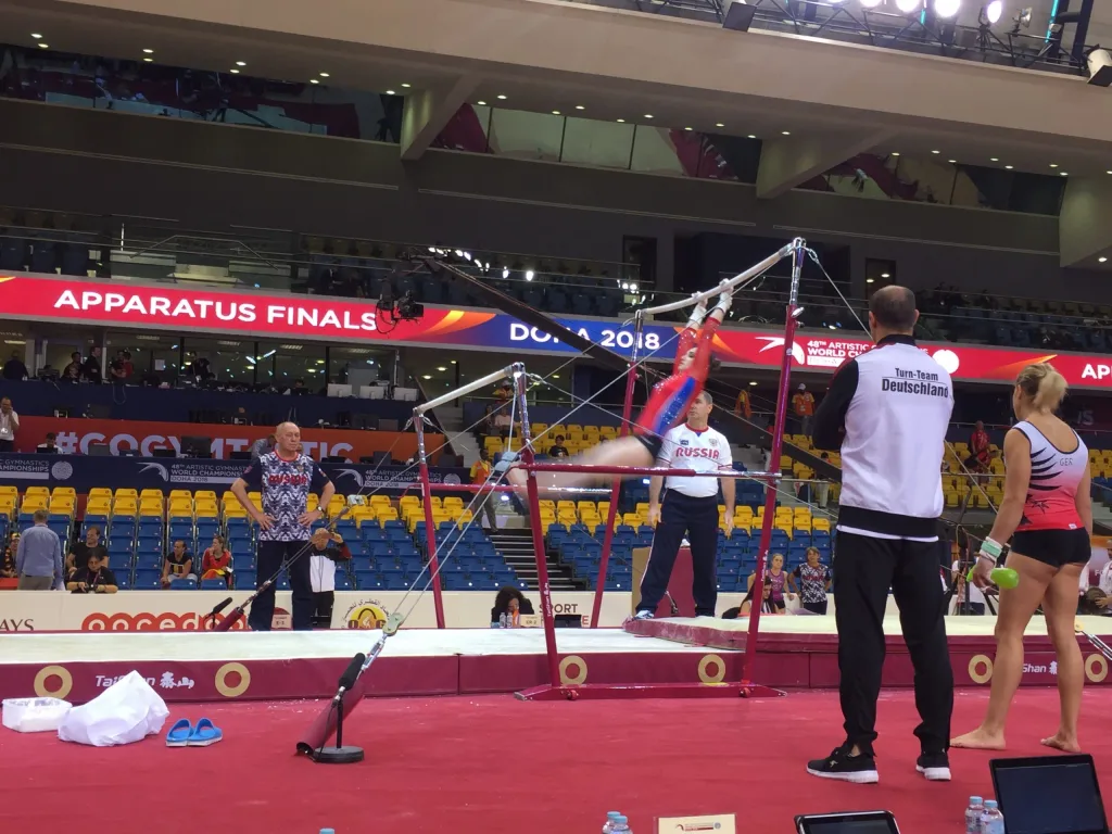 Gymnast on red uneven bars with fibreglass rails and beech veneer, coaches nearby labeled Turn-Team Deutschland.