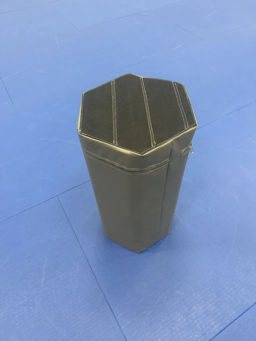 Hexagonal padded foam training table wrapped in gray PVC with a black stitched top and side zipper, standing on a blue mat.