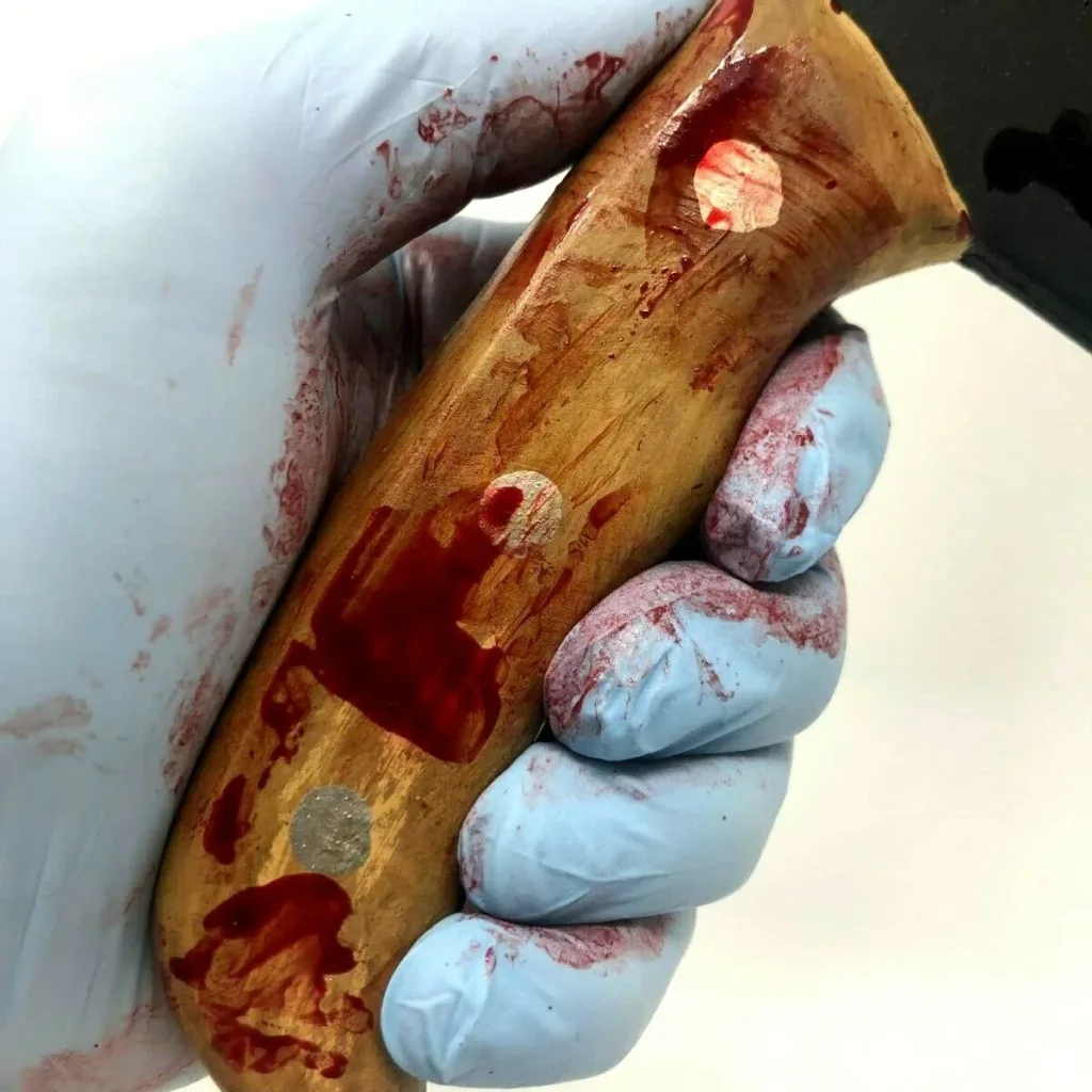 Blue-gloved hand holding a PU foam small meat cleaver training prop with wood-look handle stained with red fake blood.