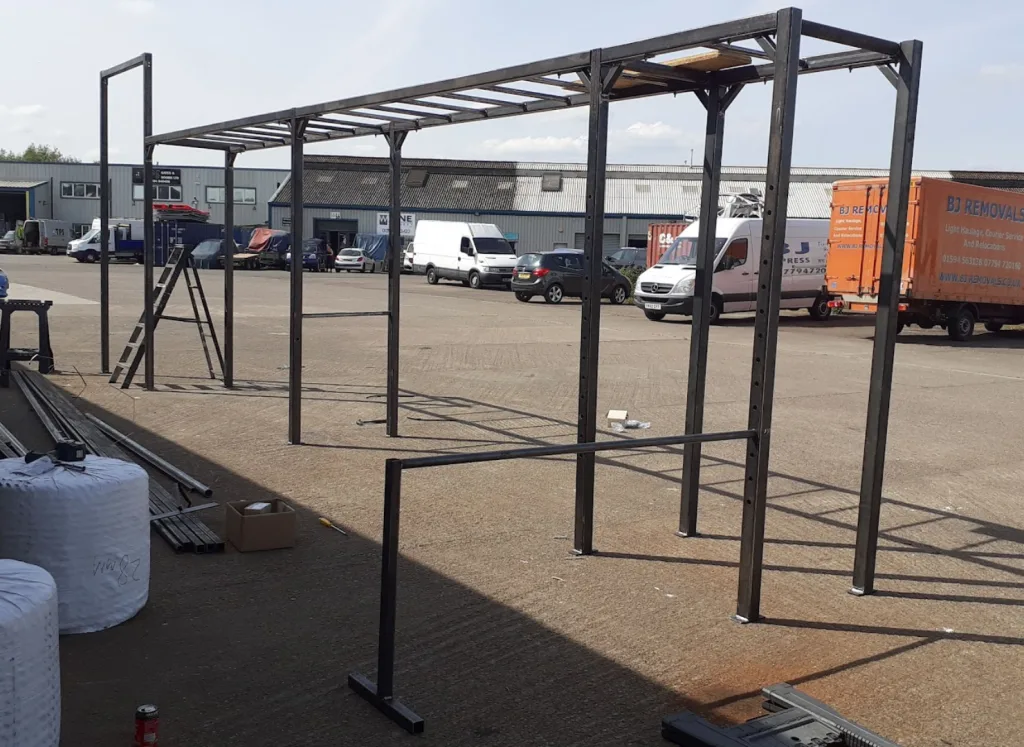 Powder-coated steel rig with uprights, monkey-bar top and lower bar, set outdoors by vans and a truck labeled BJ REMOVALS.