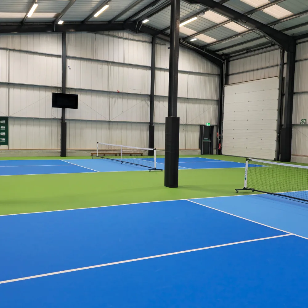 Indoor pickleball courts with blue playing surfaces, green surrounds, nets, and black PVC-covered foam padding on vertical posts.