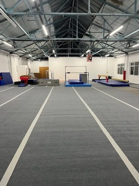 Large indoor gym hall with light grey carpet roll-out mats laid in lanes toward gymnastics apparatus at the far end.
