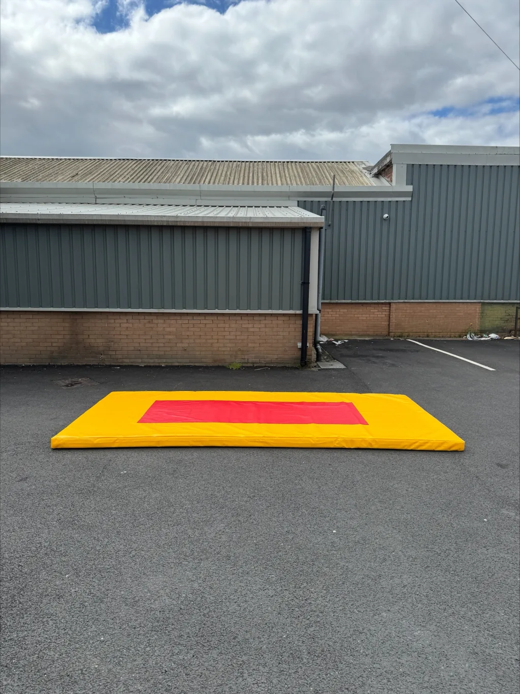 Bright yellow gymnastics mat with a central red target zone lying on asphalt outside a gray industrial building under cloudy sky.