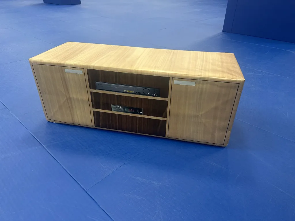 High-density PU foam TV stand with lifelike wood-print PVC cover and central shelves with printed electronics, on blue matting.