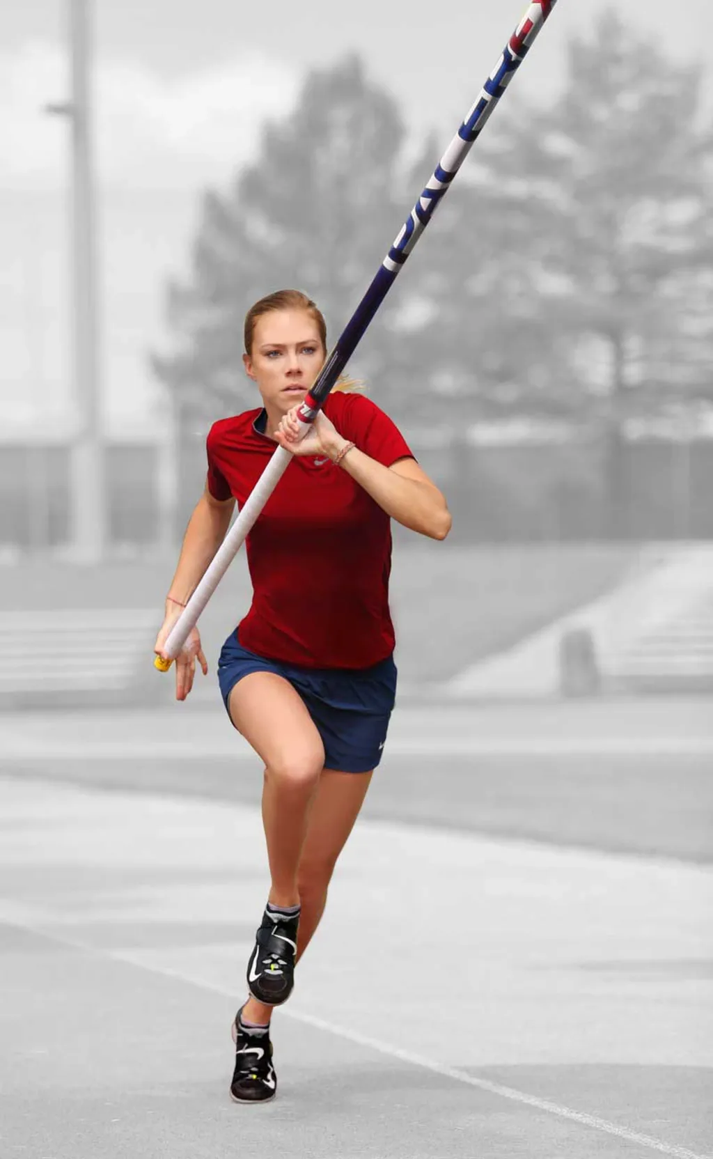 Female pole vaulter in a red shirt and navy shorts running on a track while holding a long vaulting pole angled upward.