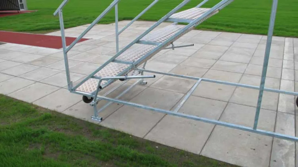 Galvanised steel and aluminium 8-step judges stand on wheels positioned on paving beside grass and a red track.