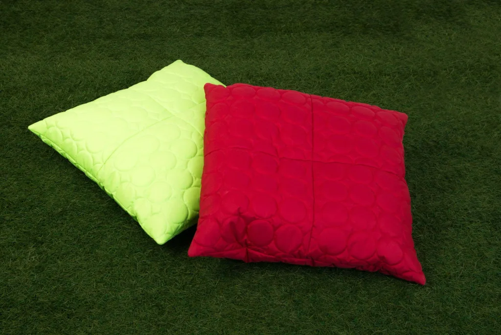 Two padded child reading cushions, one neon yellow and one red, quilted with circular stitching, lying on green grass.