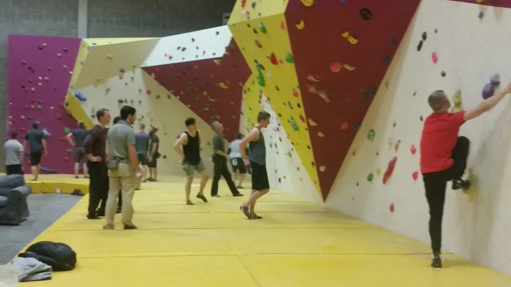 Yellow PVC bouldering mats cover the floor beneath angled indoor climbing walls with several people bouldering and observing.