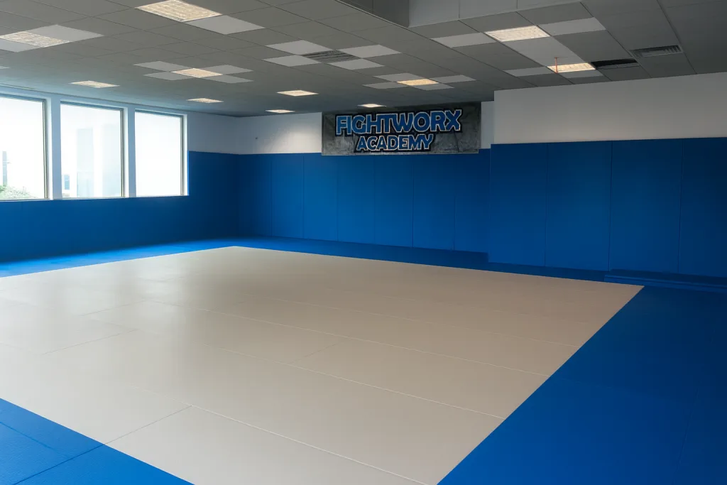 Empty martial arts room with royal blue wall padding and a white central mat, banner reading FIGHTWORX ACADEMY on the far wall.
