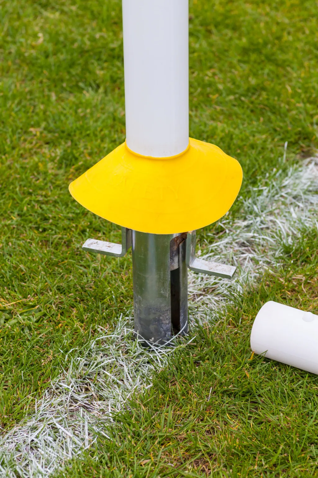 White rugby post seated in a metal sleeve with a yellow collar embossed "SAFETY" on green grass by a painted white line.