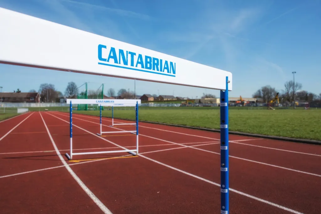 White training hurdle with the text 'CANTABRIAN' on the UPVC top board and blue fixed legs standing on a red running track under a blue sky.