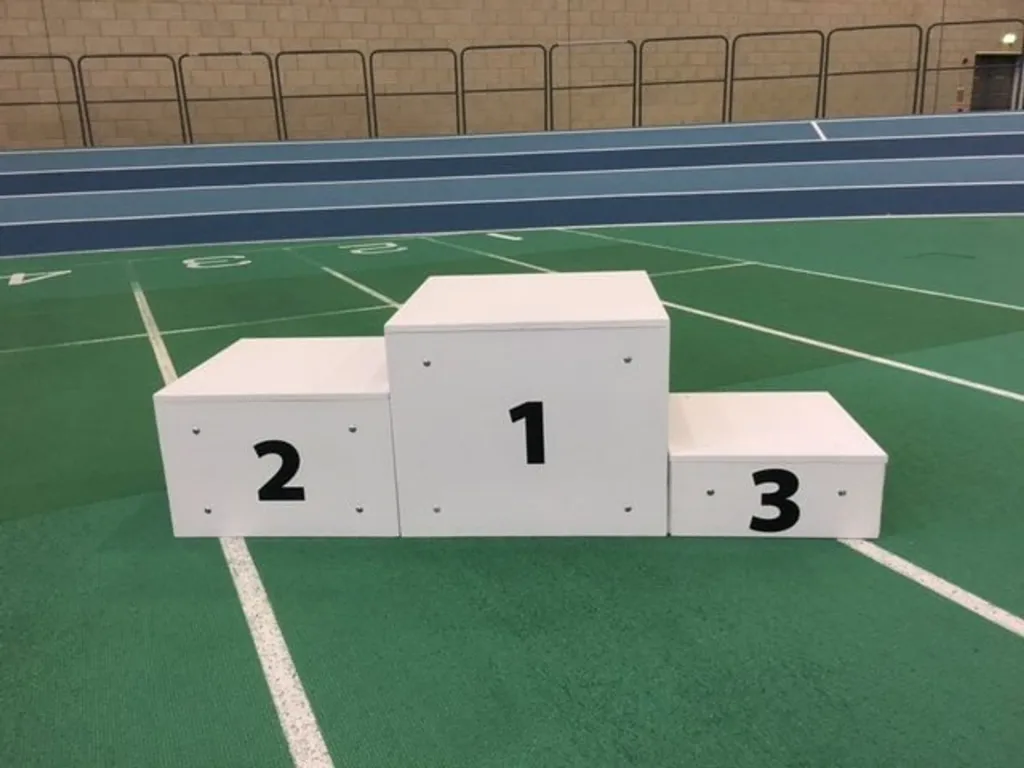 White three-step winners podium with black numbers '1', '2', '3' placed on a green indoor athletics track.