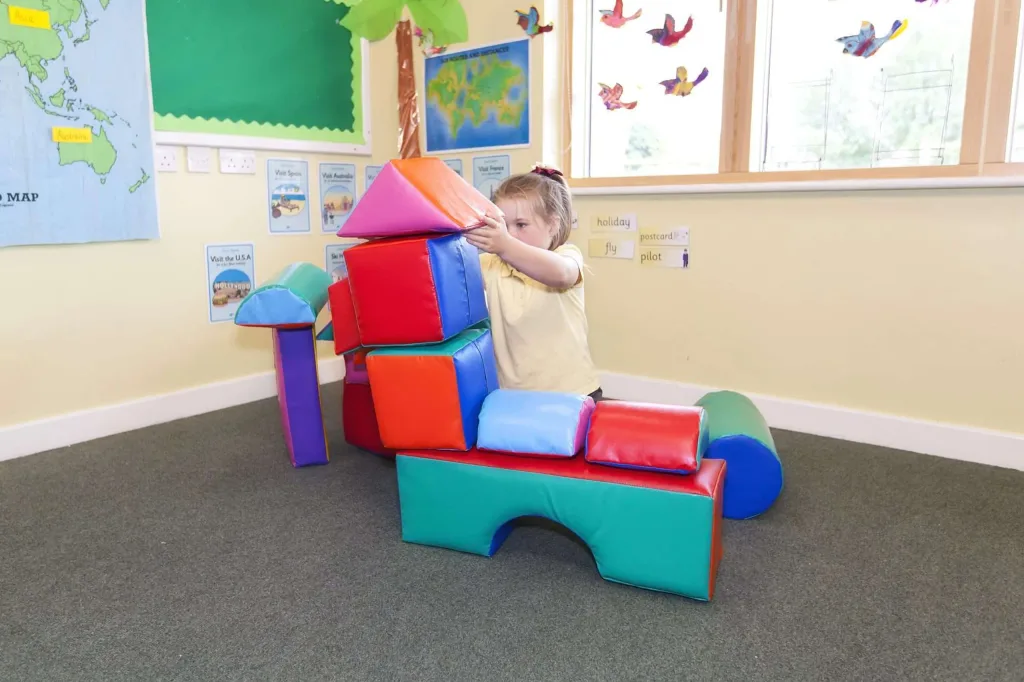 Child stacking multicolored building shapes in a classroom, wall labels read "holiday" "postcard" "fly" "pilot".