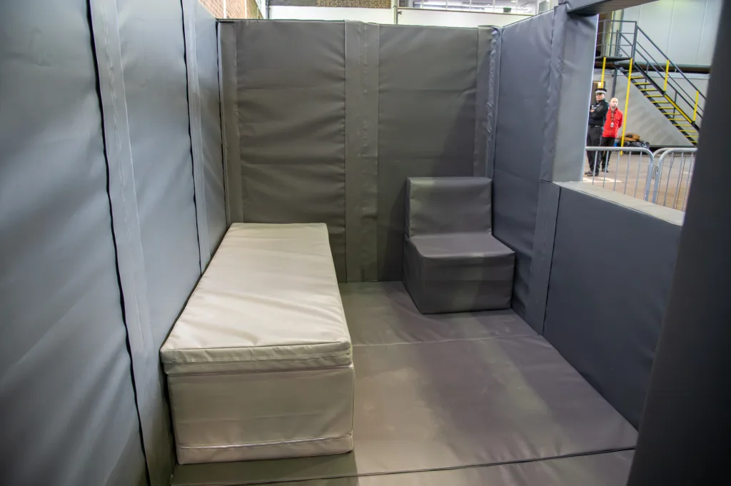 Foam-padded custody cell with heavy-duty PVC walls and floor, a padded bench along the left wall and a small padded seat in the corner.