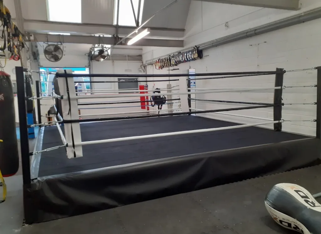 Platform training ring with 500mm raised deck, black canvas and skirt, alternating white and black ropes and steel posts inside a gym.