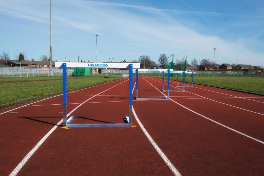 Row of steel competition hurdles reading CANTABRIAN placed on a red running track under a blue sky.