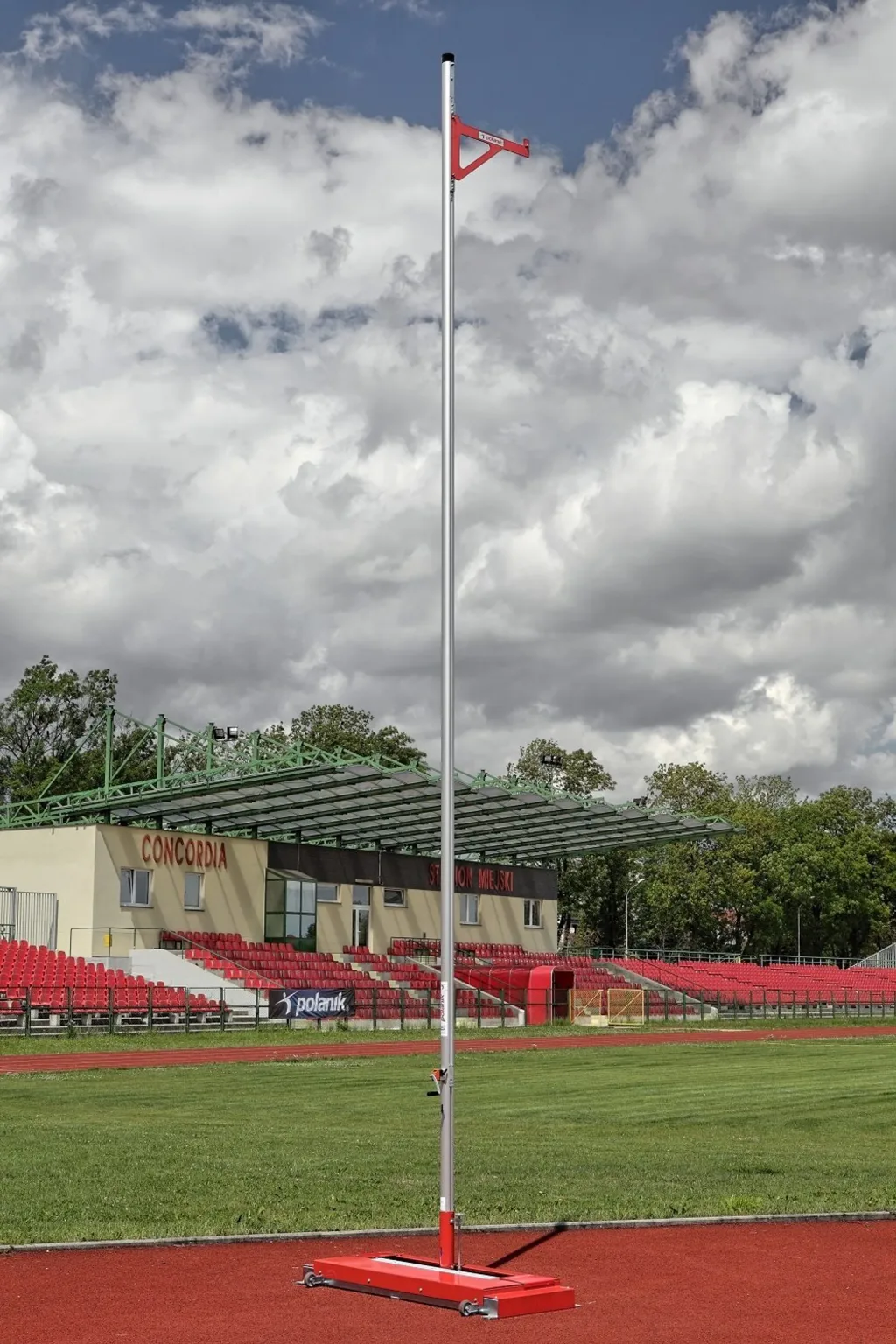 Pole vault upright with anodized aluminium post and galvanized steel base on a red track in front of bleachers reading CONCORDIA.