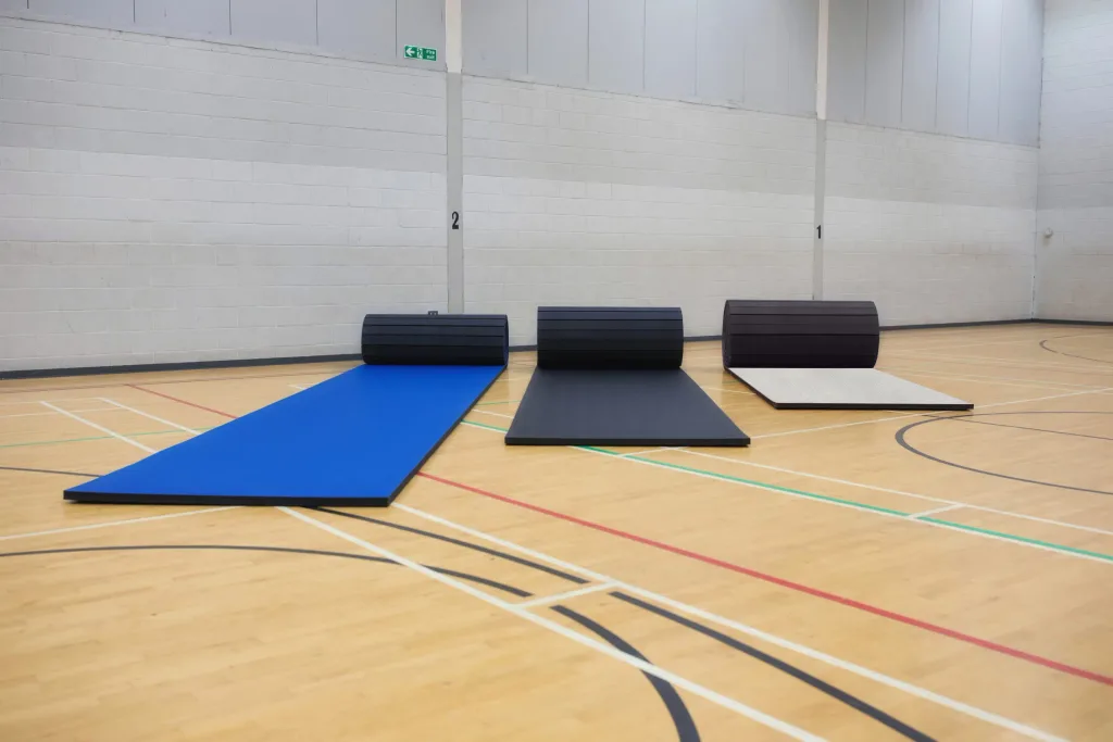 Three roll-out gym mats—blue, black, and grey—partly rolled at the far end of a wooden sports hall floor with wall columns marked "2" and "1".