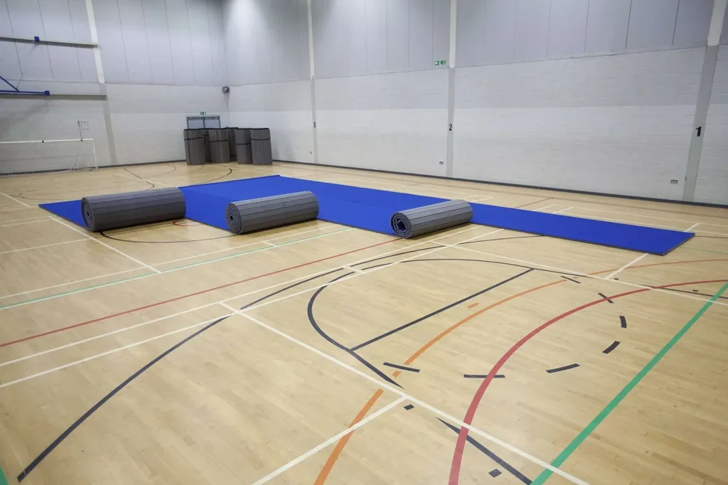 Blue carpet roll-out mat partially unrolled on a gym floor with three light grey rolled mats nearby.