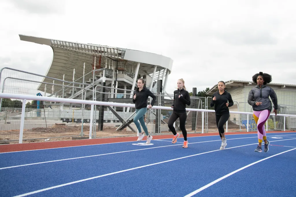 Four women jogging on a blue track in front of metal crowd safety barriers and a partially constructed stadium.
