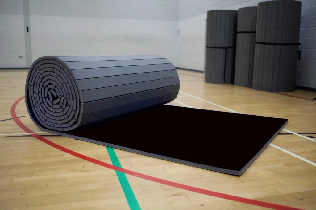 Light grey roll-out carpet mat with black surface partially unrolled on a gym floor near a wall marked 1, with three rolled mats stacked behind.