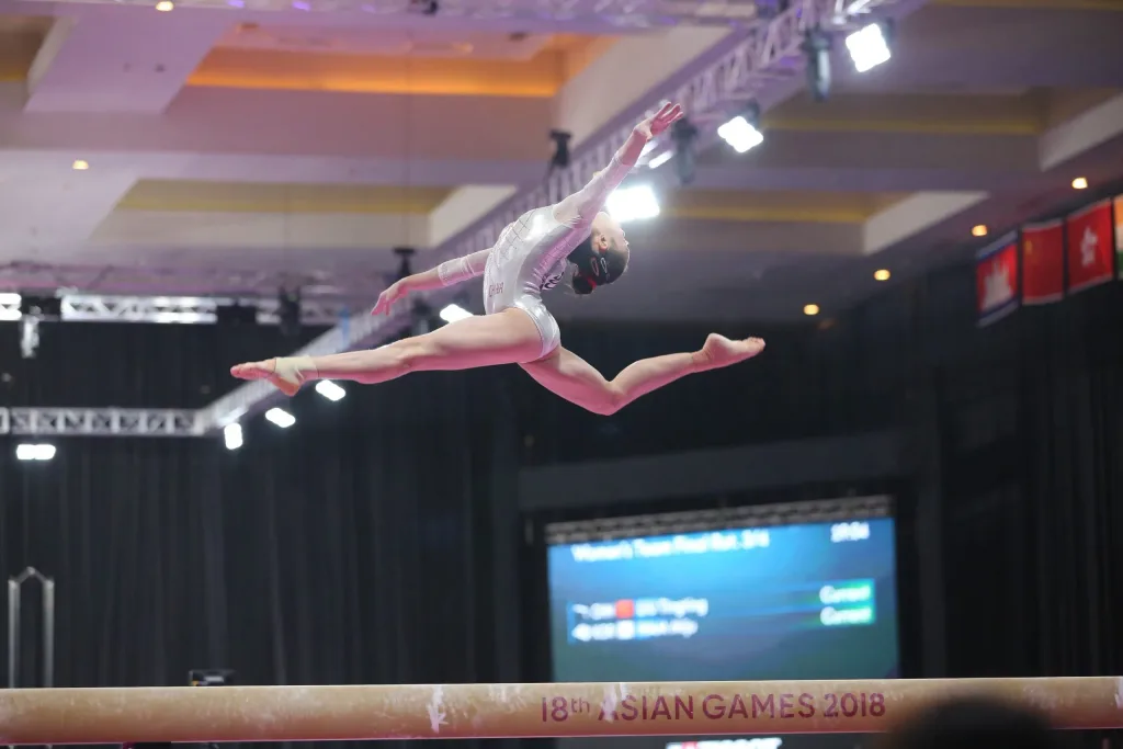 Gymnast in silver leotard performing a split leap above a red balance beam labeled "18th ASIAN GAMES 2018".