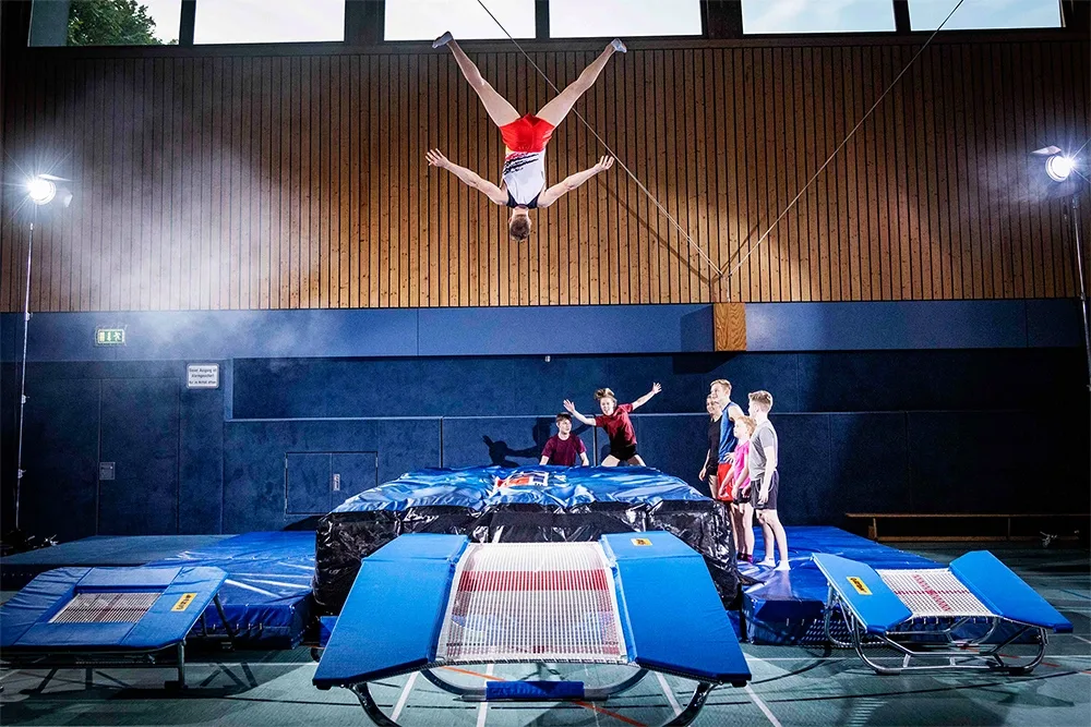 Gymnast flipped upside down above blue trampolines as five spotters watch on a gym mat in front of a wooden-paneled wall.